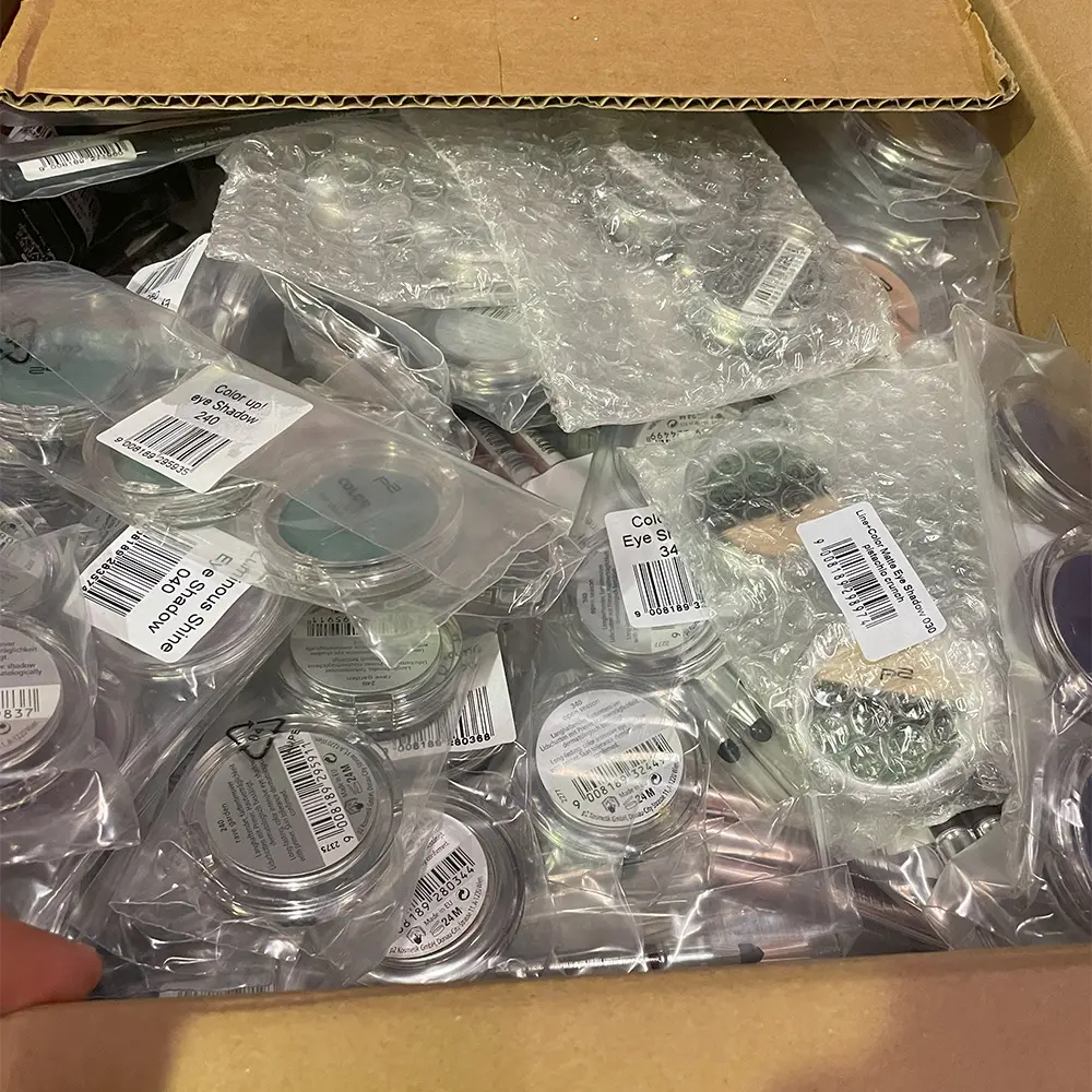 Open cardboard box filled with bubble-wrapped and plastic-packaged small cosmetic containers and makeup pencils.